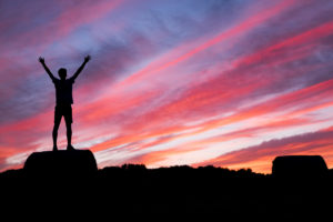 Silhouette of person standing on rocks in front of pink, purple and blue sunrise skies